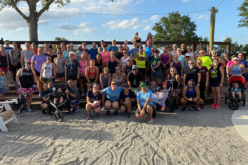 A large group of people gathered for a run club event, smiling and posing for a photo outdoors. The group is diverse, with some participants in athletic wear, sunglasses, and hats, and a few with strollers, indicating a family-friendly event. They are standing in a sandy area with string lights overhead and trees in the background, suggesting a casual, community atmosphere.