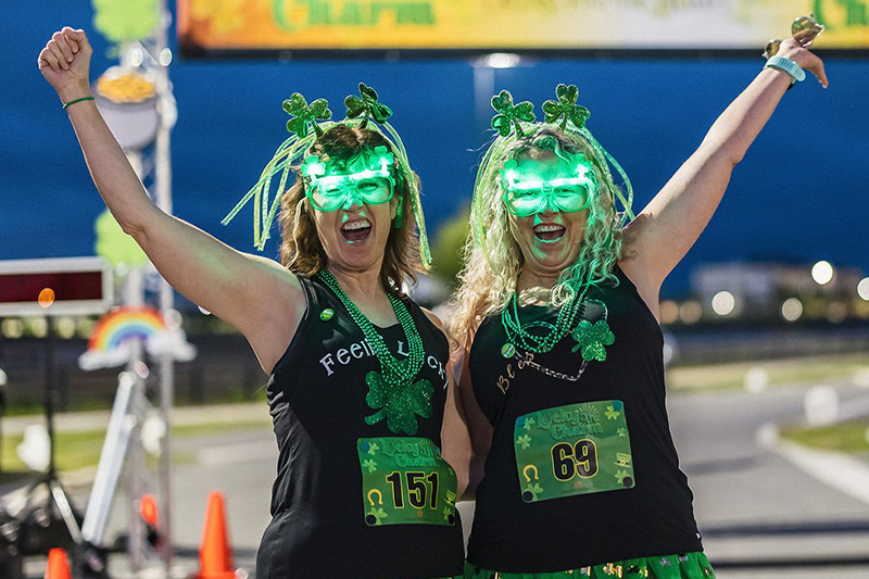 Two participants at the Lucky Charm 5k event in Hamlin, joyfully raising their arms in victory, wearing green shamrock-themed costumes, including tutus and illuminated headbands. They stand at the starting line under a banner that reads "Lucky Charm 5k," with a nighttime sky in the background.