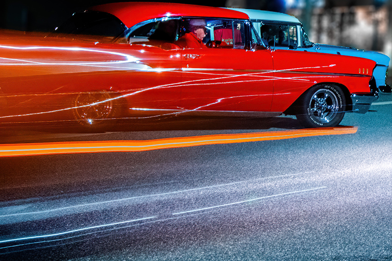 A vintage red and white classic car in motion at night, with streaks of light reflecting off its shiny surface creating a dynamic effect, indicative of speed and the vibrant energy of a cruise-in event.