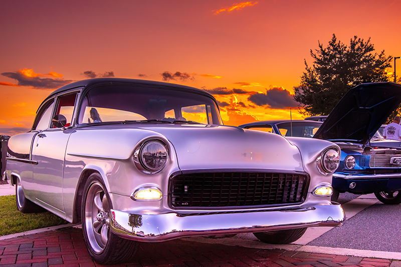 A classic silver two-door sedan from the mid-20th century is parked at a cruise-in event during sunset, with the hood open displaying the engine. The car's chrome details gleam under the vibrant hues of the dusk sky. Another vintage car with its hood open is visible in the background.