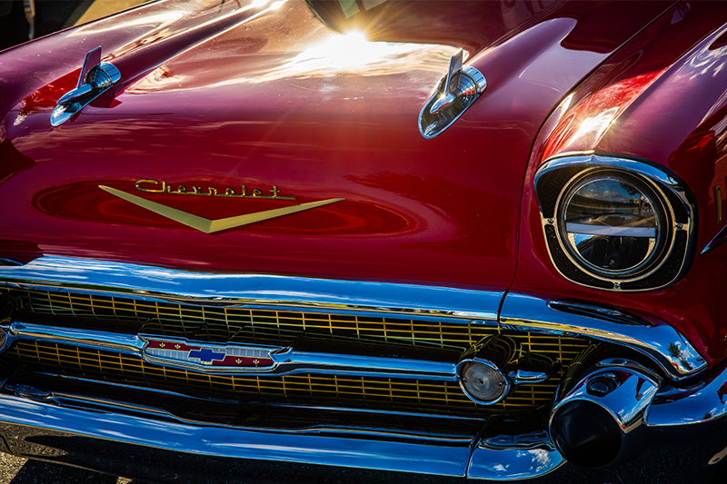 The front view of a classic Chevrolet car in a shiny red color, featuring distinctive chrome details and hood ornaments. The Chevrolet emblem is prominently displayed on the front of the hood, with the sunlight reflecting off the polished surface.