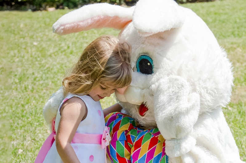A young girl in a pastel Easter dress gently hugs a person in a large, fluffy white Easter Bunny costume. The bunny is sitting down on a sunny grass field, and the girl is leaning her head affectionately against the bunny's chest.