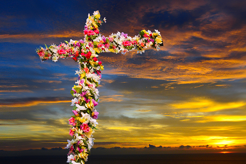 A cross covered with multicolored flowers is silhouetted against a dramatic sunrise sky with clouds streaked in shades of yellow, orange, and blue.