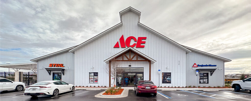 Front view of a new Ace Hardware store with a large, red 'ACE' sign above the entrance. The white building features the STIHL and Benjamin Moore logos, with cars parked in front under a cloudy sky.