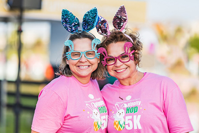 Two women wearing pink "Hippity Hop 5K" event t-shirts smiling at the camera, sporting festive Easter-themed glasses with attached bunny ears in sparkling blue and pink sequins, at an outdoor 5K run.
