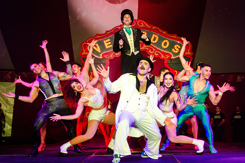 A lively performance by the Venardos Circus cast on stage. Performers are dressed in colorful, vibrant costumes, with a mix of sequins, leotards, and formal circus attire. The ringmaster stands at the back, wearing a top hat and black suit with tails, while other performers in front strike dynamic, joyful poses. A bold red marquee with "Venardos" written in gold letters is displayed in the background, enhancing the festive, theatrical atmosphere. The scene captures the excitement and energy of this Broadway-style circus performance.