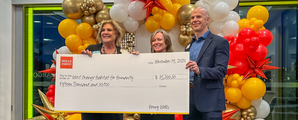 A celebratory photo of three individuals holding a large ceremonial check during the grand opening of Wells Fargo’s new Hamlin branch. The check is made out to "West Orange Habitat for Humanity" for $15,000 and dated November 19, 2024. The backdrop features a festive arrangement of gold, red, and white balloons with star accents, creating a vibrant and cheerful atmosphere. The donation supports efforts to construct homes in Winter Garden, Florida.