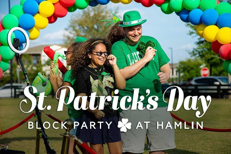 A joyful St. Patrick’s Day celebration at Hamlin featuring a diverse group of people dressed in festive green attire. A man wearing a green leprechaun hat and a green "Lucky" shirt is smiling while holding a glittery shamrock decoration. A young girl in the foreground, wearing a black shirt with a green design, is laughing and dancing with friends. She wears fun shamrock-shaped sunglasses and green bead necklaces. Behind them, a colorful balloon arch made of green, yellow, blue, and red balloons marks the entrance to the event. Other attendees in green clothing can be seen celebrating in the background under a bright blue sky.