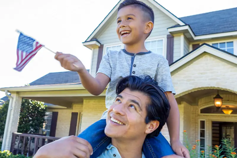 A father smiles up at his young son riding on his shoulders, waving a small American flag in front of a suburban home in Horizon West, Florida, celebrating the Hamlin All-American Flag Parade.
