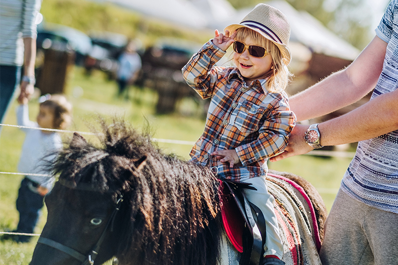 A young child wearing sunglasses and a straw hat sits atop a miniature pony, tipping their hat with a smile while an adult steadies them during a sunny outdoor community event.